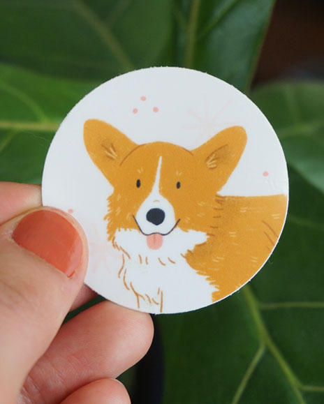 A hand holds a corgi sticker in front of some greenery