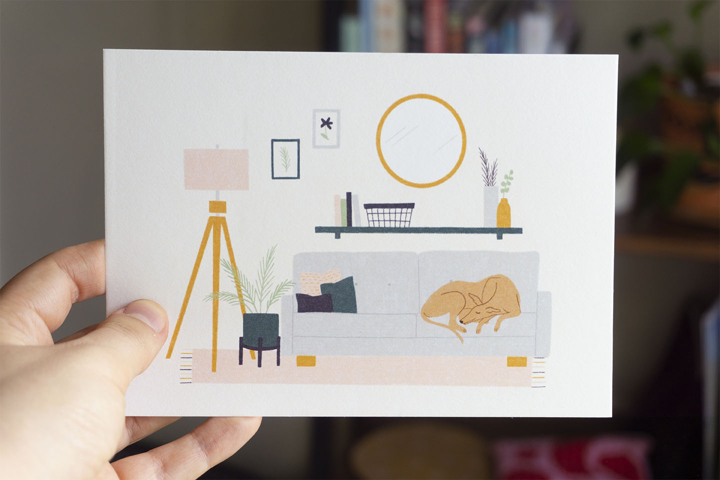 A hand holds an art print featuring a greyhound napping in a living room