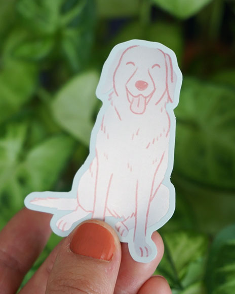 A hand holds a labrador sticker in front of some greenery