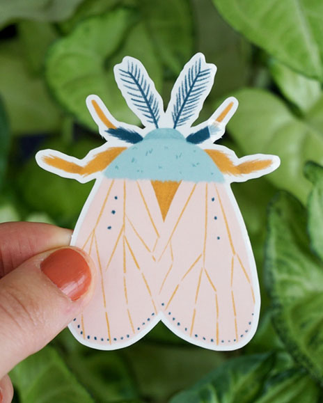 A hand holds a moth sticker in front of some greenery
