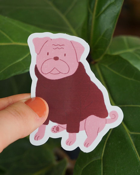 A hand holds a Pug sticker in front of some greenery