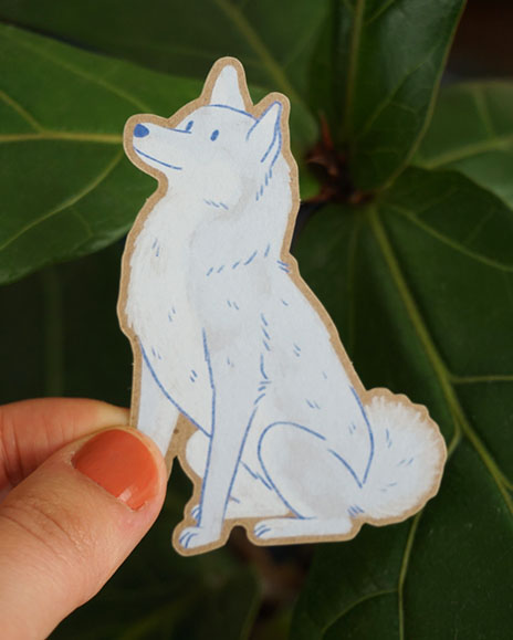 A hand holds a Shiba Inu sticker in front of some greenery