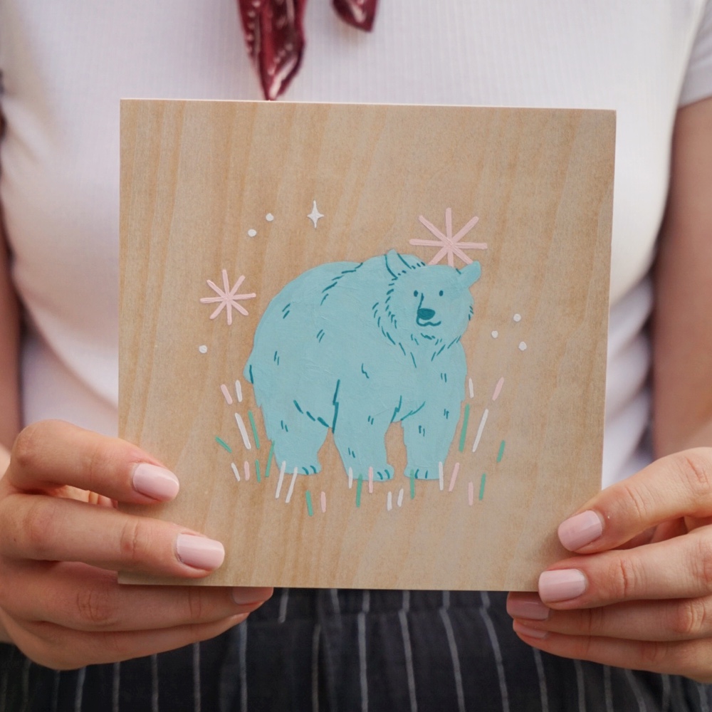 Someone holds a gouache painting of a grizzly bear on a 6x6 wood panel