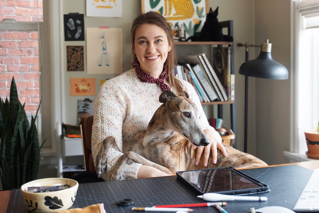Kaila sits at her desk with her greyhound sitting on her lap