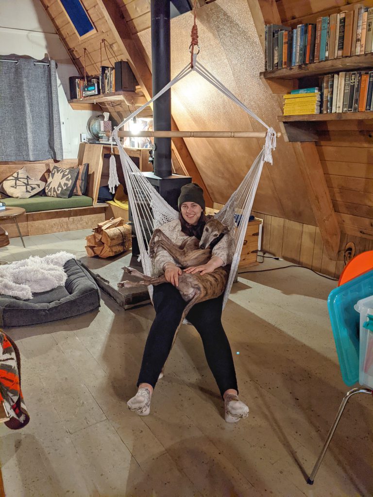 Kaila sits in a woven swinging chair with her greyhound, Greer, on her lap