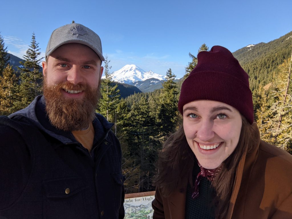Ian (a white man in his mid-twenties with a big red beard and grey cap) and Kaila (a woman in her mid twenties with long brown hair and a red hat) pose for a selfie with Mt. Rainier behind them