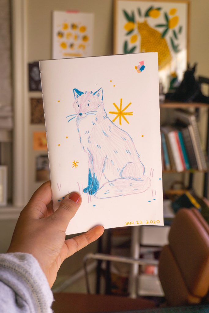 A hand holds out a colored pencil drawing of a blue and purple fox