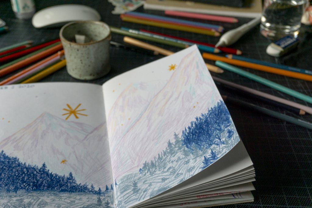 a closeup view of a sketchbook filled with a landscape of purple mountains and blue trees. pencil crayons and a small ceramic jar full of pencil shavings are in the background