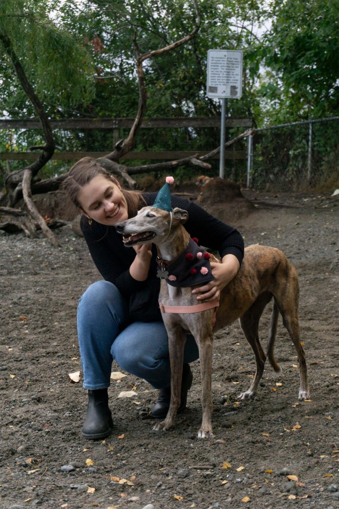 Kaila, a brown ahired white woman, holds her smiling greyhound who is wearing a blue party hat with a pom pom