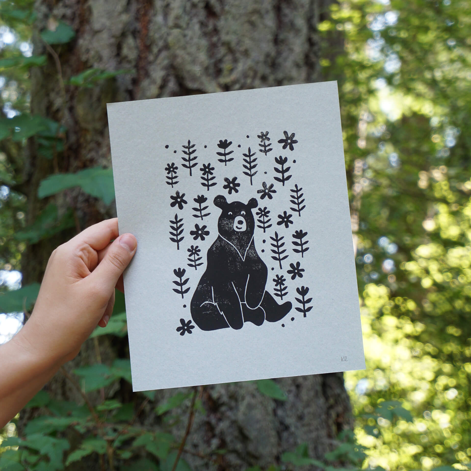 A hand holds up a linocut print of a bear sitting among flowers