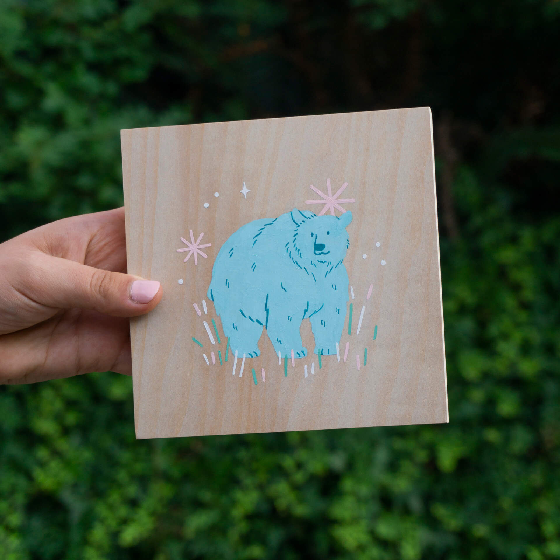 A hand holds a light wood panel that has a blue bear painted on it