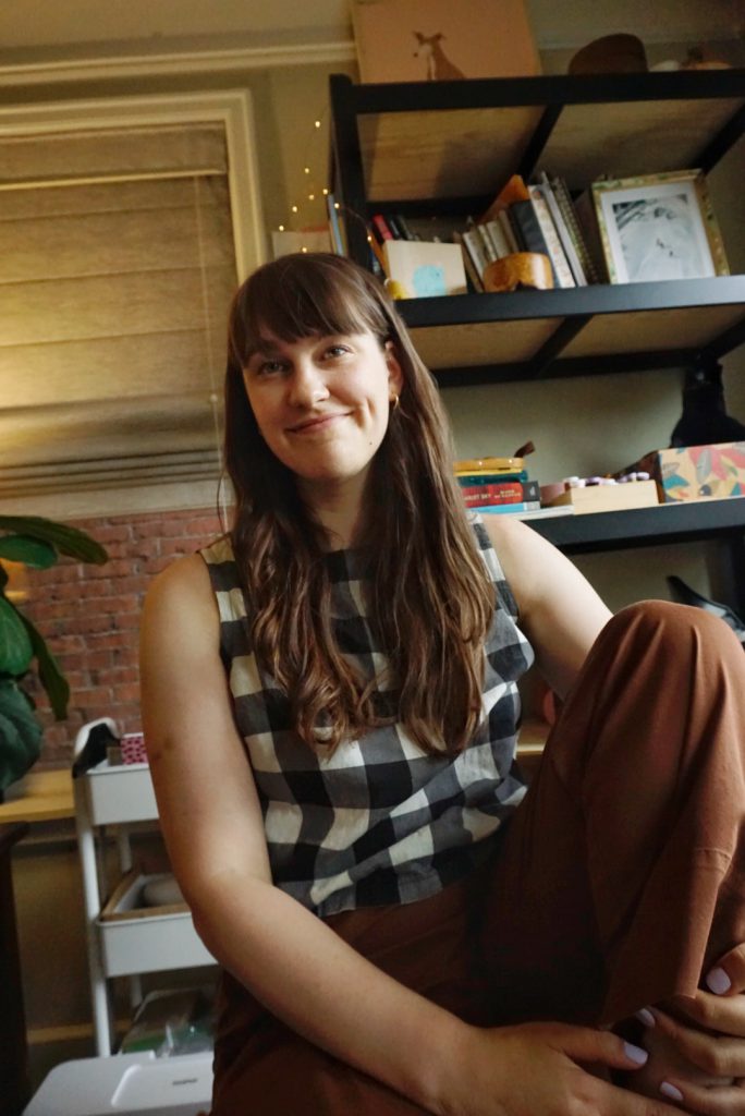 Kaila, a 30 yr old white woman with long brown hair sits casually and smiles at the camera, holding one knee that's bent up onto the chair she sits on.