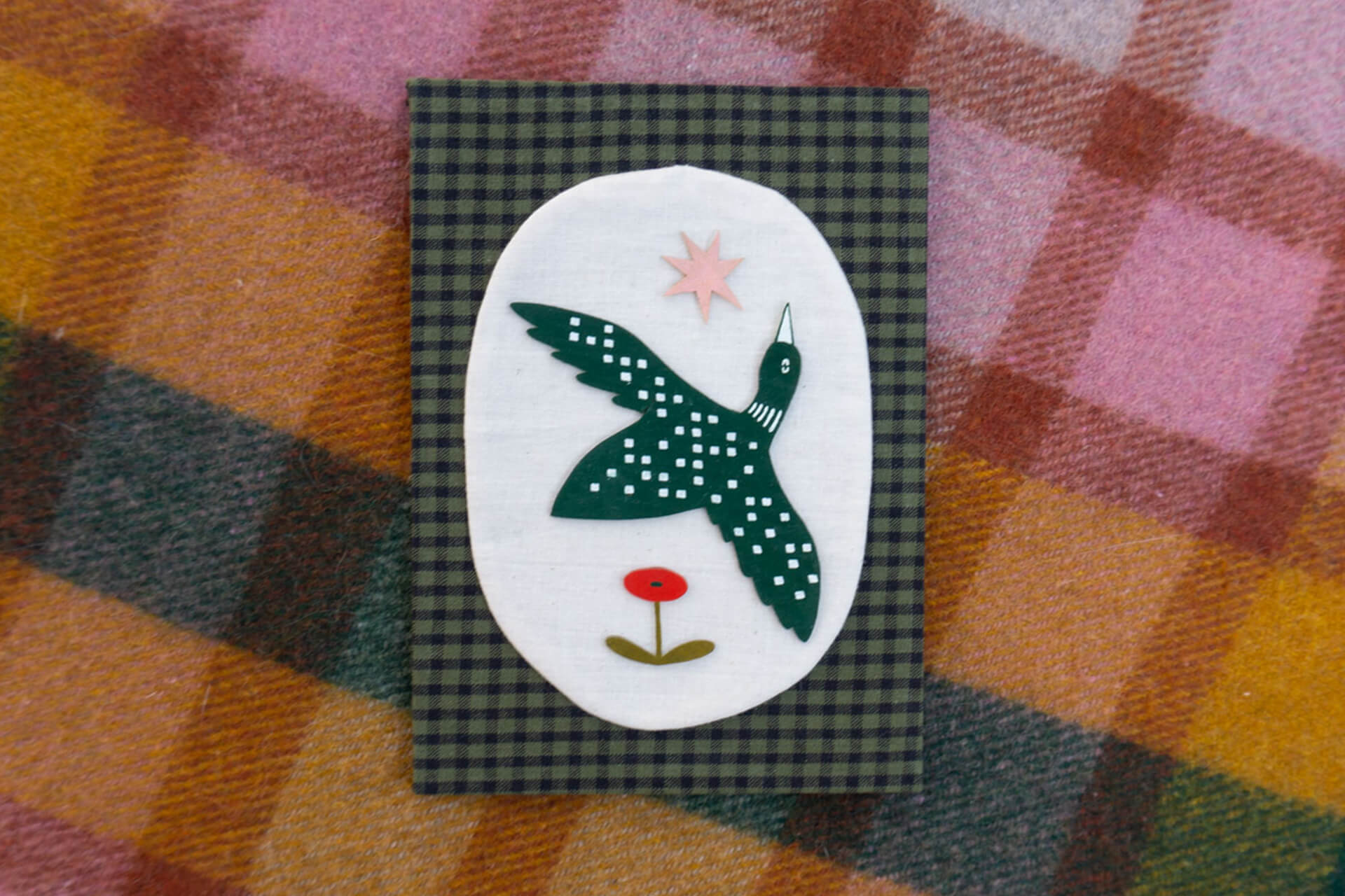 A mixed media artwork of a loon flying between a star and a poppy. It sits on a beige linen oval on top of a green gingham fabric background